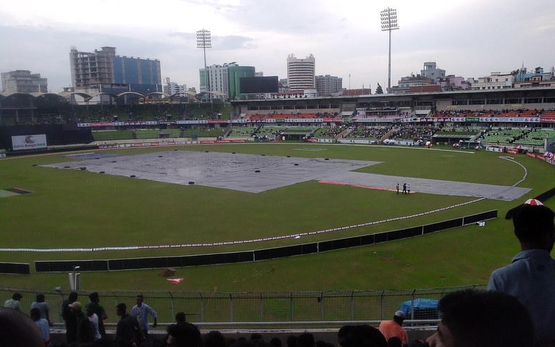 Rain covers spread over the pitch under grey skies as play is stopped for rain at a South Asian cricket ground. Photo: Xkalponik / Wikimedia Commons (CC BY-SA 4.0)