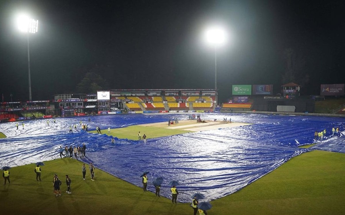 Rain covers spread over the TU Ground outfield under floodlights as ground staff shelter under umbrellas during rain at Kirtipur