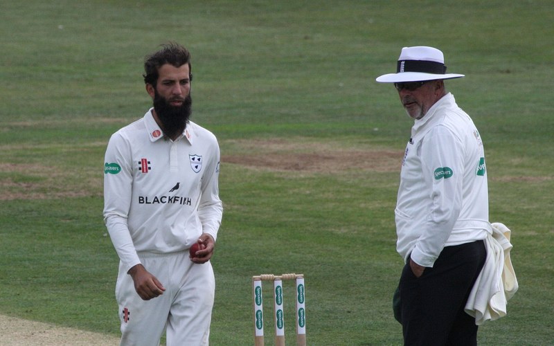 Moeen Ali in whites holding a cricket ball on the field. Photo: Dave Morton / Wikimedia Commons (Public domain)